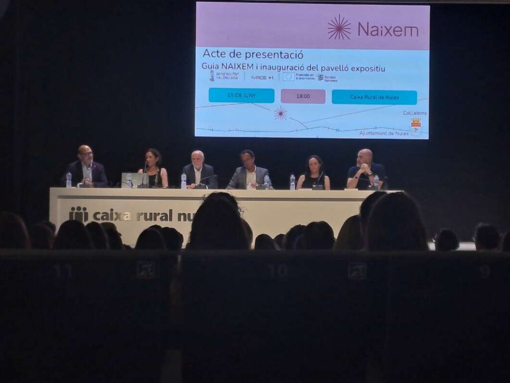 Panel de seis ponentes sentados detrás de una mesa larga en un escenario, con una pantalla de presentación detrás.