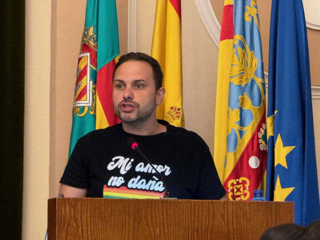 Man speaking at a wooden podium with a microphone, colorful flags behind him in a formal setting.