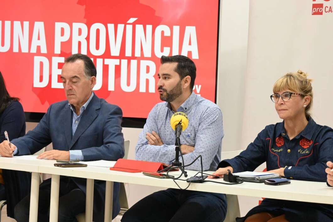 Three people sit at a conference table during a press briefing: a man in a blue suit writes, another man speaks into a yellow microphone, and a woman with glasses listens as papers and devices lie on the table, with a red banner in the background.