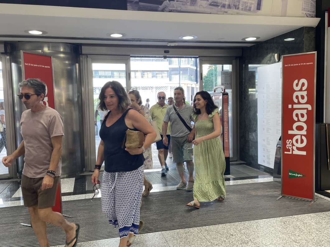 People exit a modern lobby through glass doors; a woman in a black top and patterned pants leads the group, while others follow behind. Red Las Palmas signs flank the doorway on the right.
