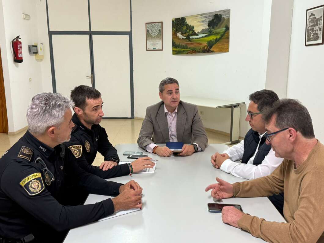 Six men sit around a table in a meeting room; two police officers on the left, three civilians on the right, center man in gray suit relevant to discussion.