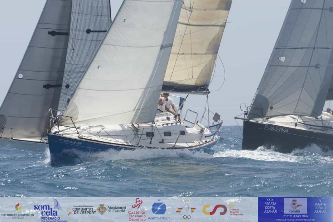 Sailing race with multiple yachts cutting through blue water, sails full and crews at the helm, sponsor banners along the shore.