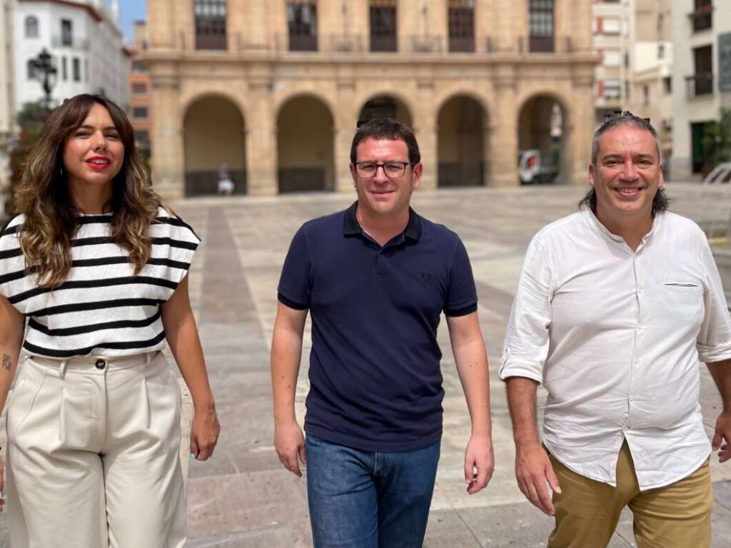 Three adults walking toward the camera in a sunny square, with a stone-arch building behind them.