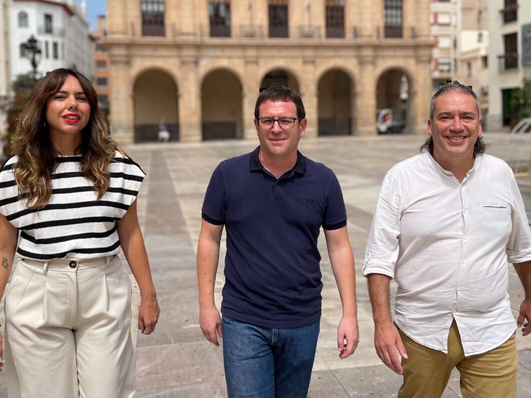 Three adults walking toward the camera in a sunny square, with a stone-arch building behind them.