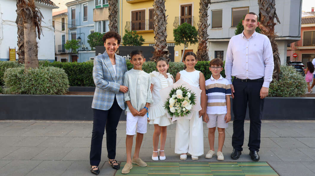 Family posing in a town plaza: two adults with four children in white outfits; a girl holds a bouquet of flowers.