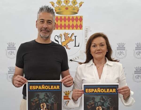 Two adults hold identical posters for a Spanish-language show, standing in front of a coat-of-arms backdrop.