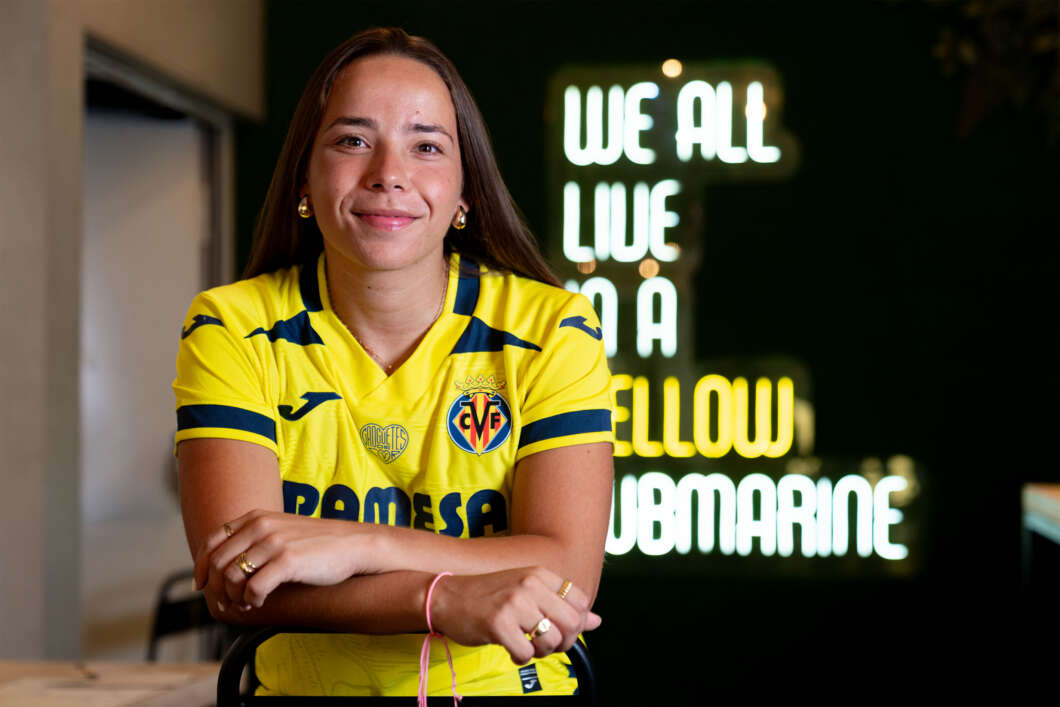Smiling young woman in a yellow soccer jersey sits with arms crossed at a table, neon sign glowing in the background.