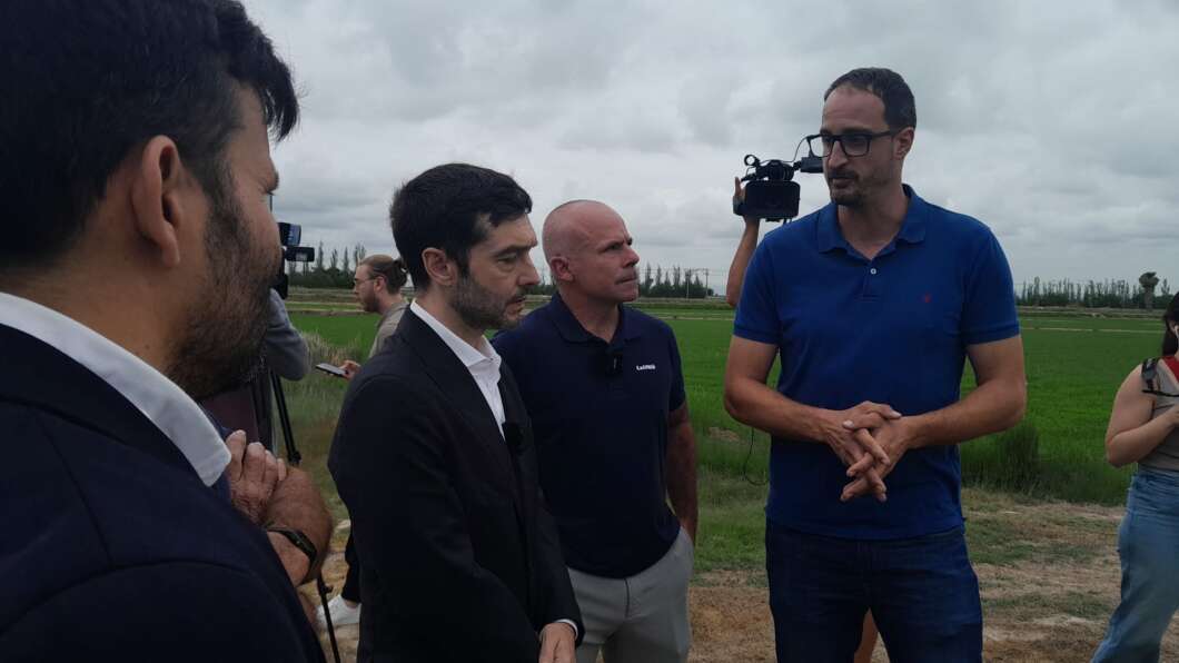 Group of men in a rural field chatting, with a tall man in a blue polo speaking while others listen and a cameraman records nearby.