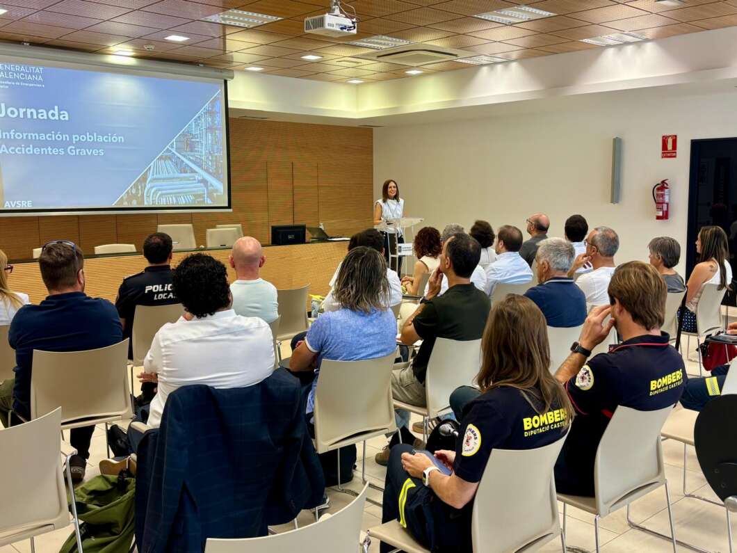 Speaker at a lectern presenting to an audience in a conference room; projection screen shows a blue slide with 'Jornada' and safety topics; some attendees wear uniforms in the foreground
