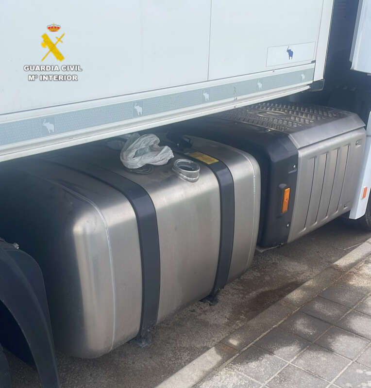 Side view of a large metal fuel tank mounted under a white truck, with a yellow filler cap and a plastic bag on top.