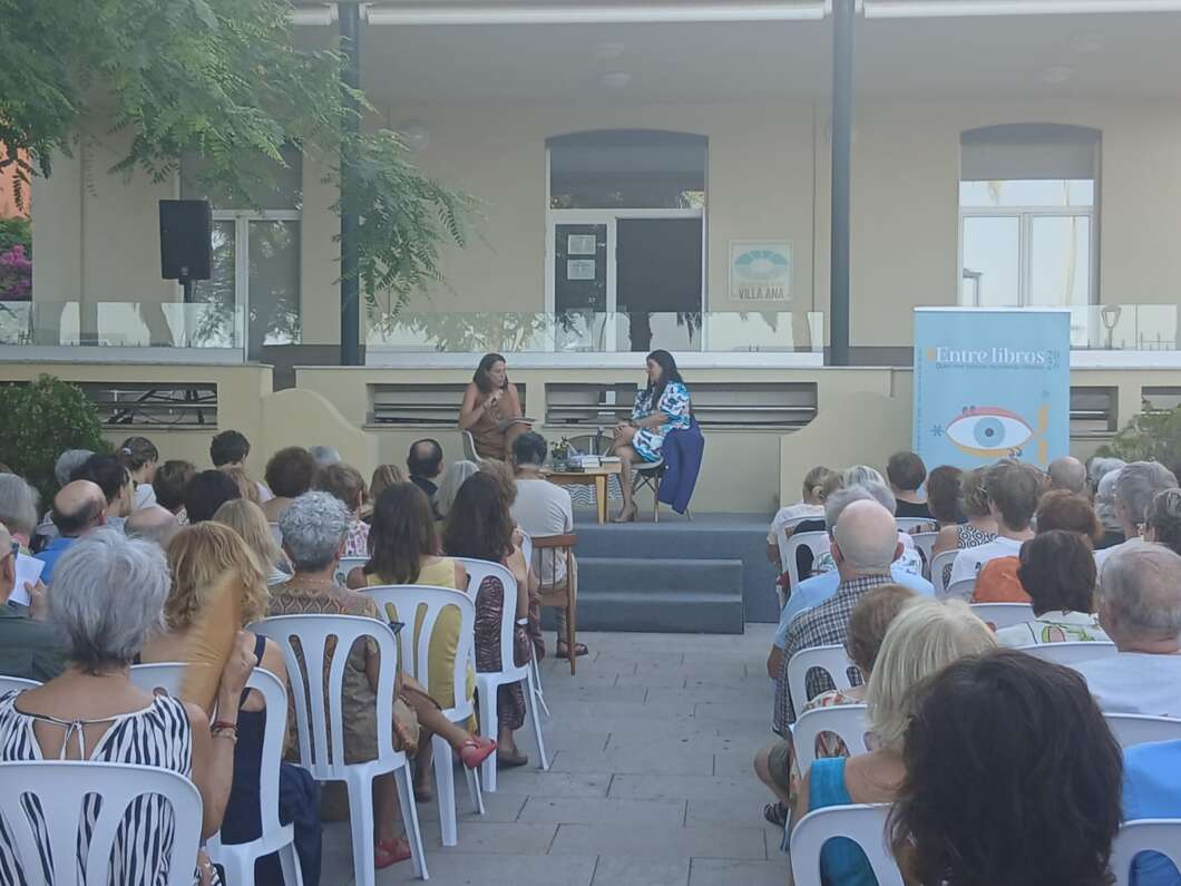Outdoor book talk with two female speakers on a stage, audience seated in white plastic chairs under trees, banner reading 'Entre libros'.