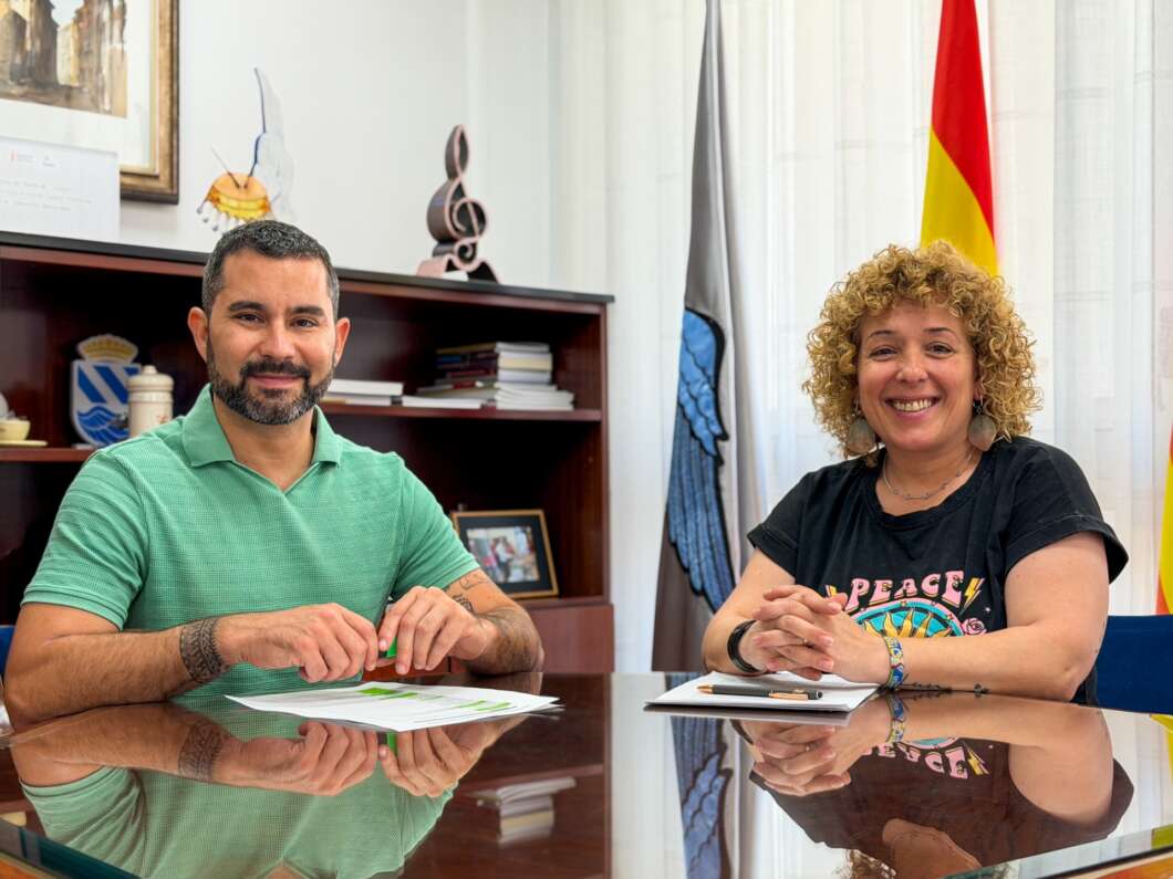 Two people sit at a glass conference table, smiling, signing documents in a bright office environment with flags in the background.