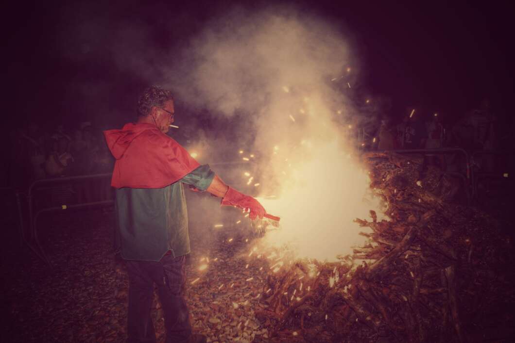 Man in a red-green jacket tending a large bonfire at night, sparks flying around him.