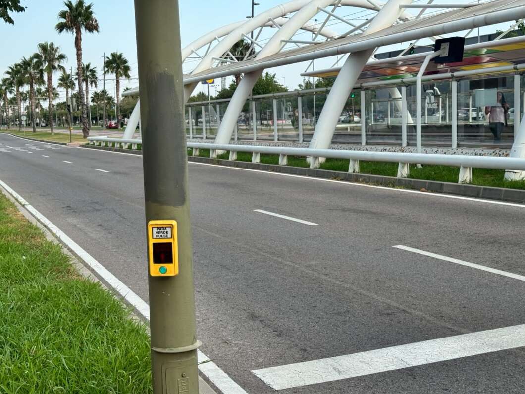 Yellow pedestrian crossing button mounted on a street pole beside a multi-lane road and a modern glass transit shelter with palm trees in the background.