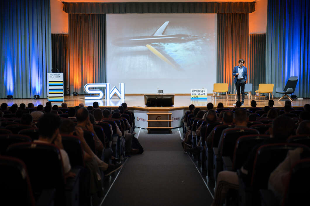 Audience in a dim auditorium watching a presenter on stage with a large screen showing a jet in flight and colorful stage lighting.