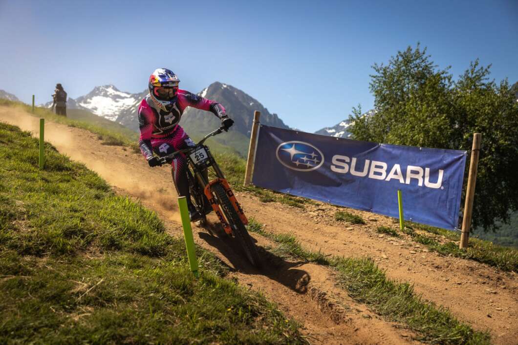 Cyclist in pink gear rides a downhill dirt trail with a Subaru banner and snowy mountains in the distance.