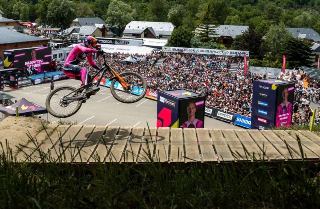 Mountain biker in bright pink gear launches off a wooden ramp at a crowded outdoor stadium event, spectators watching from stands and banners in the background.