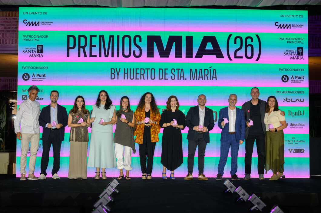 Group of award recipients on stage posing with pink trophies in front of a bright Premios MIA(26) backdrop.