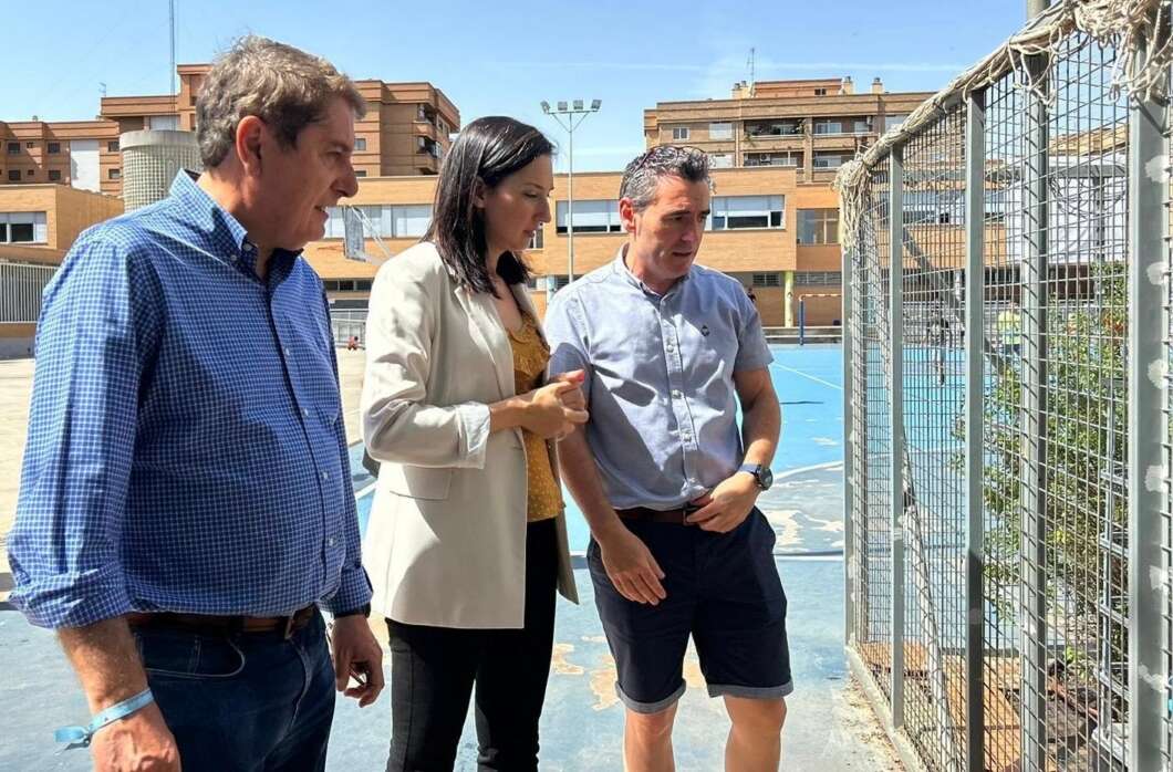 Three adults, two men and a woman, stand on a sunny blue outdoor sports court beside a tall chain-link fence, looking intently at something beyond the fence.
