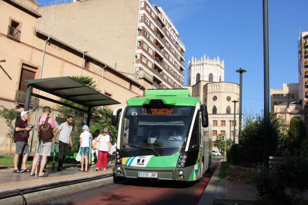 Green modern tram bus at a sunny urban stop with passengers waiting on the sidewalk.