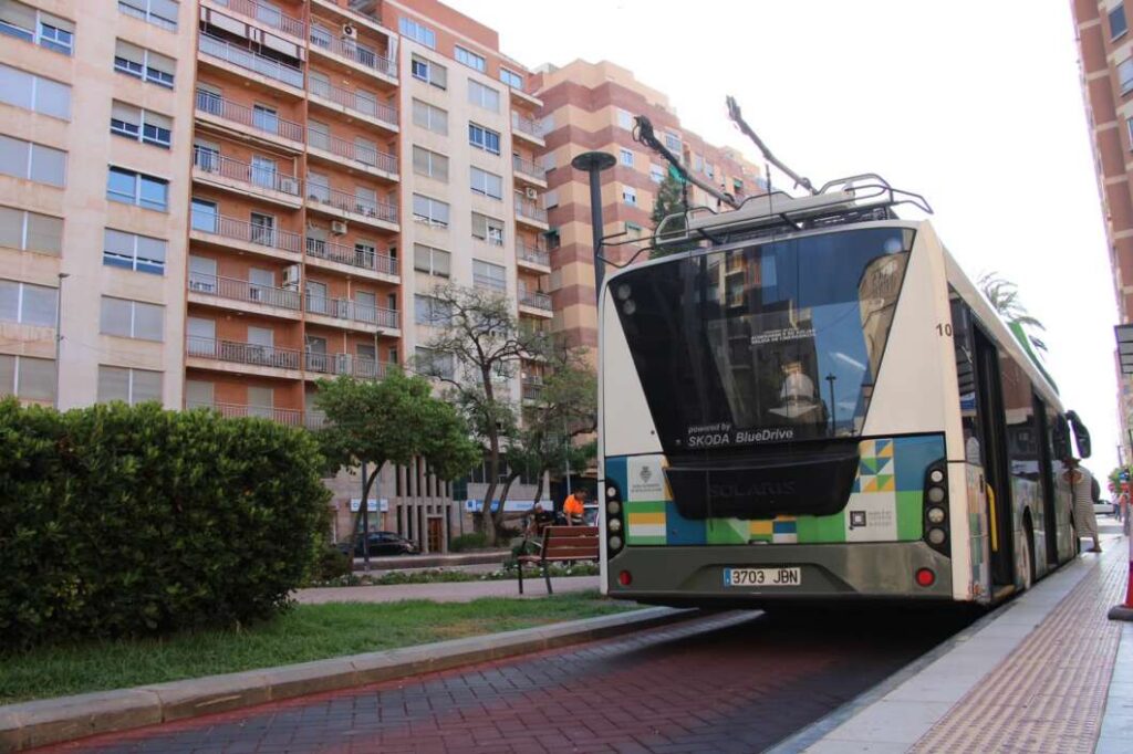 Articulated city bus at a curb in a residential area with beige apartment buildings and a small park nearby.