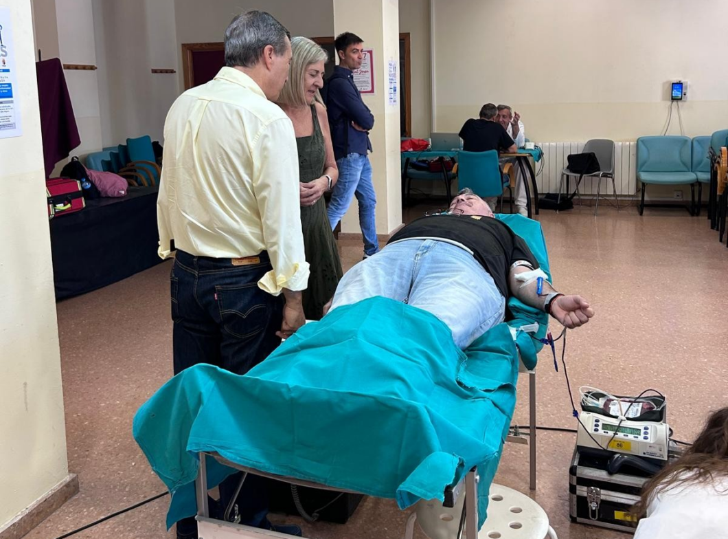Man lying on a hospital bed donating blood, IV in his arm, as others stand nearby in a clinic room.