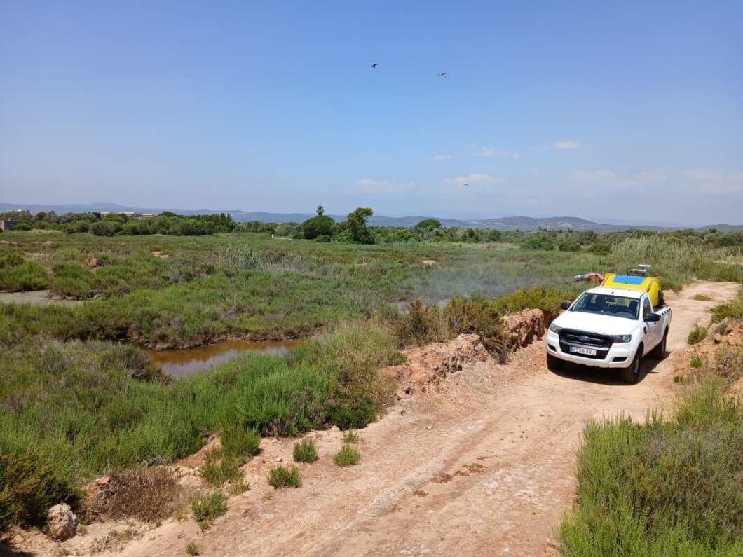 White pickup truck with yellow equipment on a dirt rural road, green scrubland and distant hills beneath a clear blue sky.
