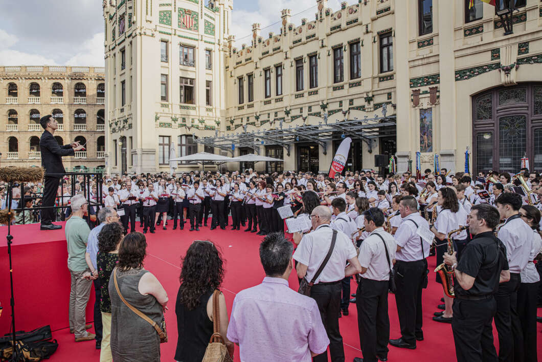Conductor leads a large choir and band performing on a red carpeted platform outdoors in front of an ornate historic building.