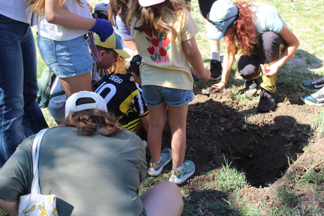 Group of kids and adults digging a hole in a grassy field on a sunny day, using gloved hands and sneakers visible around the hole.