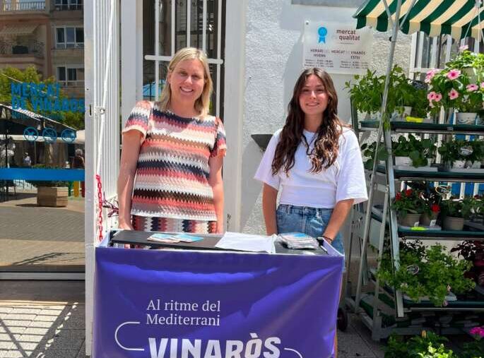 Two women stand behind a purple Vinaròs information table outdoors, with plants and a striped canopy nearby, promoting the Mediterrani event.