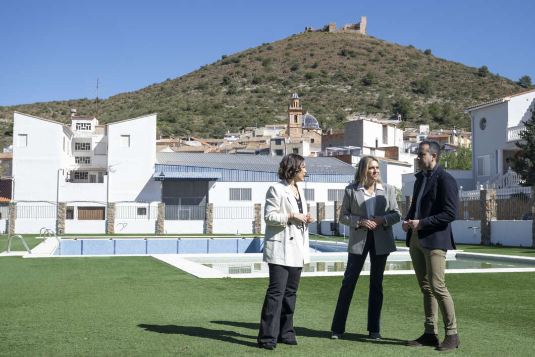 Three professionals in smart casual attire stand on a green lawn outside a residential complex, talking with a hillside and a castle ruin in the background beside a rectangular pool.