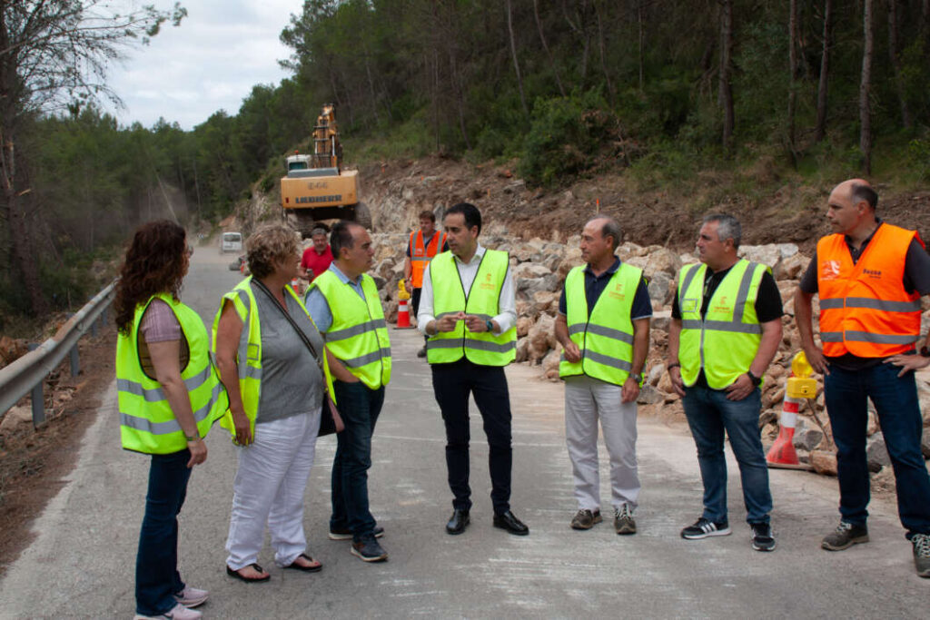 Group of construction workers and supervisors in high-visibility vests standing on a road under construction, discussing with an excavator and rock wall in the background.