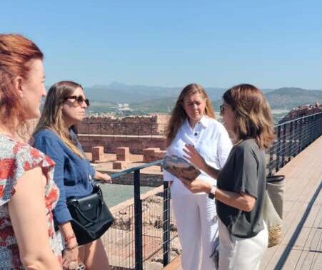 Four women chat on a sunny boardwalk with a railing, overlooking rocky terrain and distant hills; one holds a brochure.