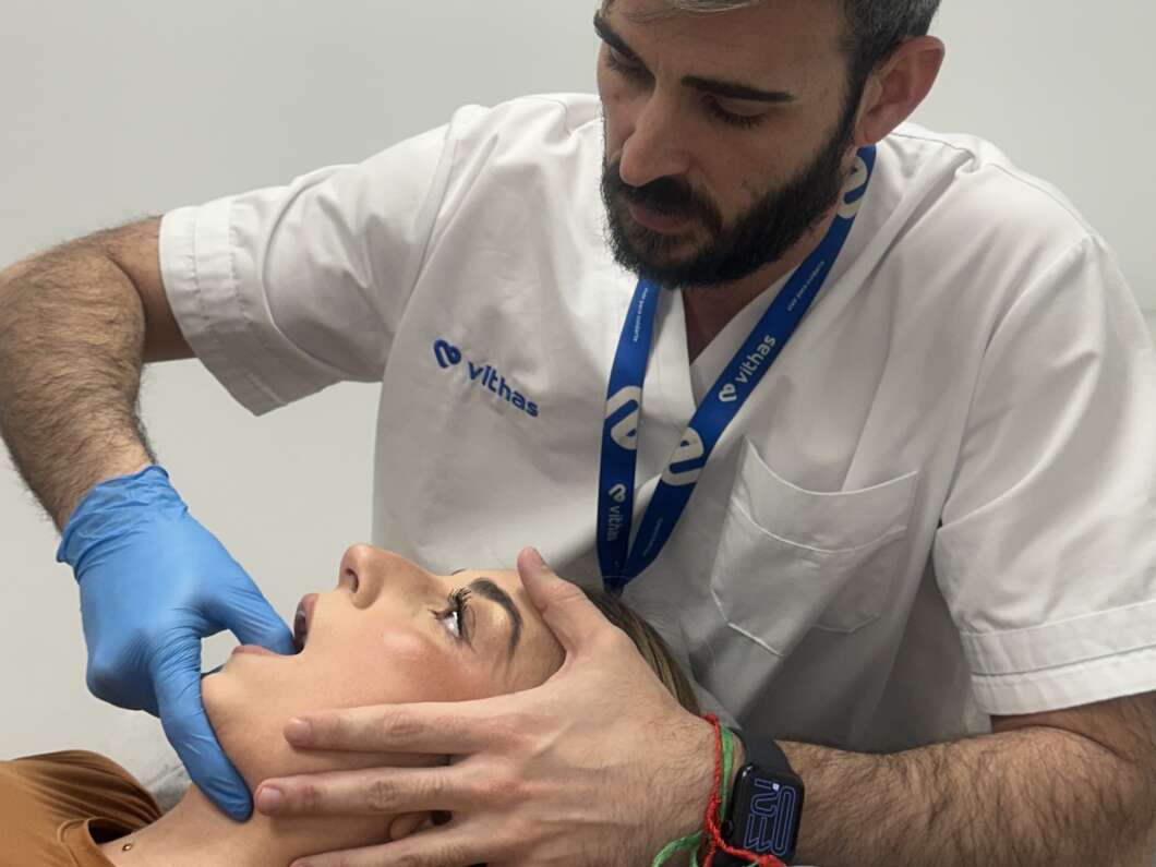 Male healthcare professional in white scrubs uses blue gloves to inspect a patient's open mouth during an examination.