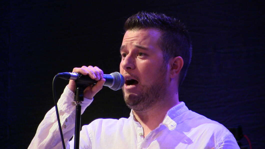 Male singer in a white shirt performing into a handheld microphone on a dark stage