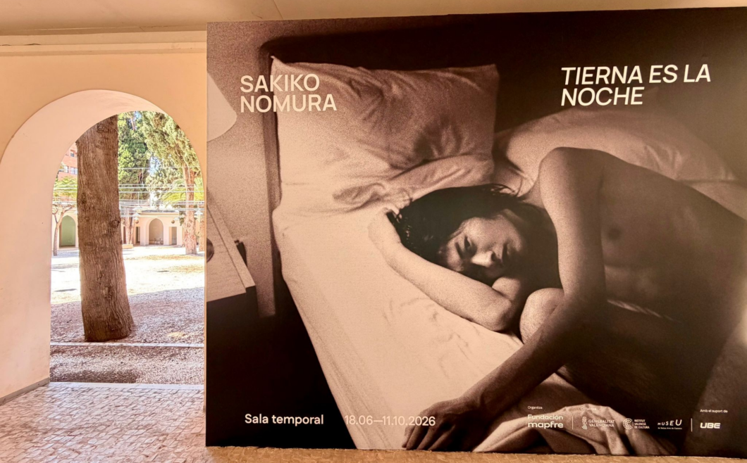 Large grayscale exhibition poster showing a topless person sleeping on a bed, with the text 'SAKIKO NOMURA' and 'TIERNA ES LA NOCHE', beside a white archway overlooking a sunlit courtyard.