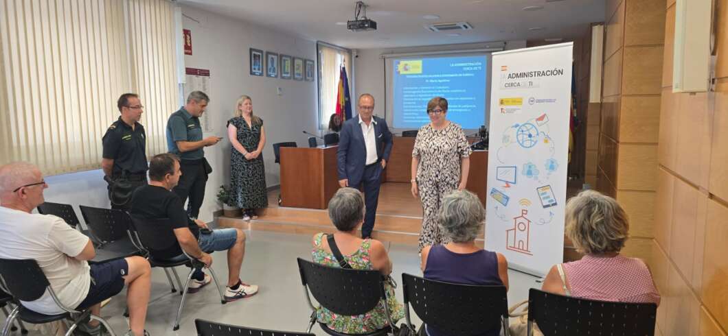 Small training room with two presenters at the front, a projector screen, and an 'Administración' banner; audience seated and watching.