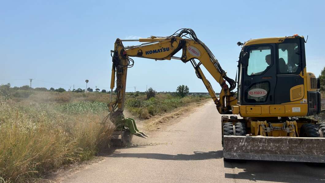 Yellow excavator on a rural road trimming tall roadside grass with a cutting attachment.