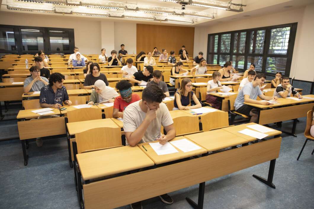 A university classroom filled with students seated at tiered desks, taking an exam with papers and pens in front of them.