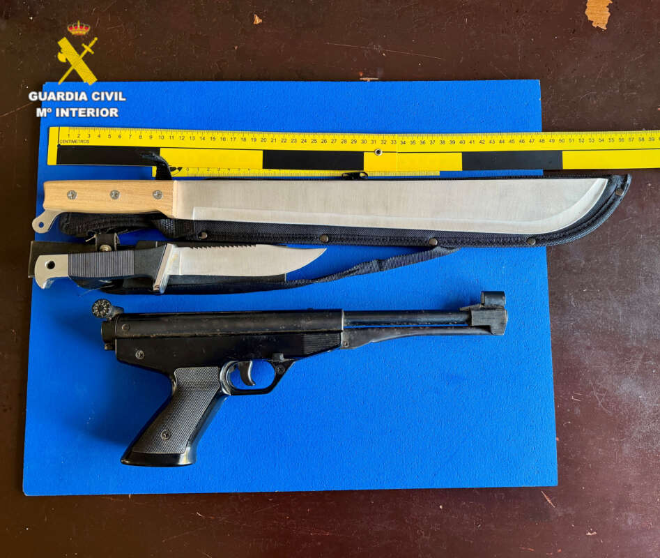 Assorted weapons laid on a blue display board with a yellow ruler; includes a long machete and a pistol.