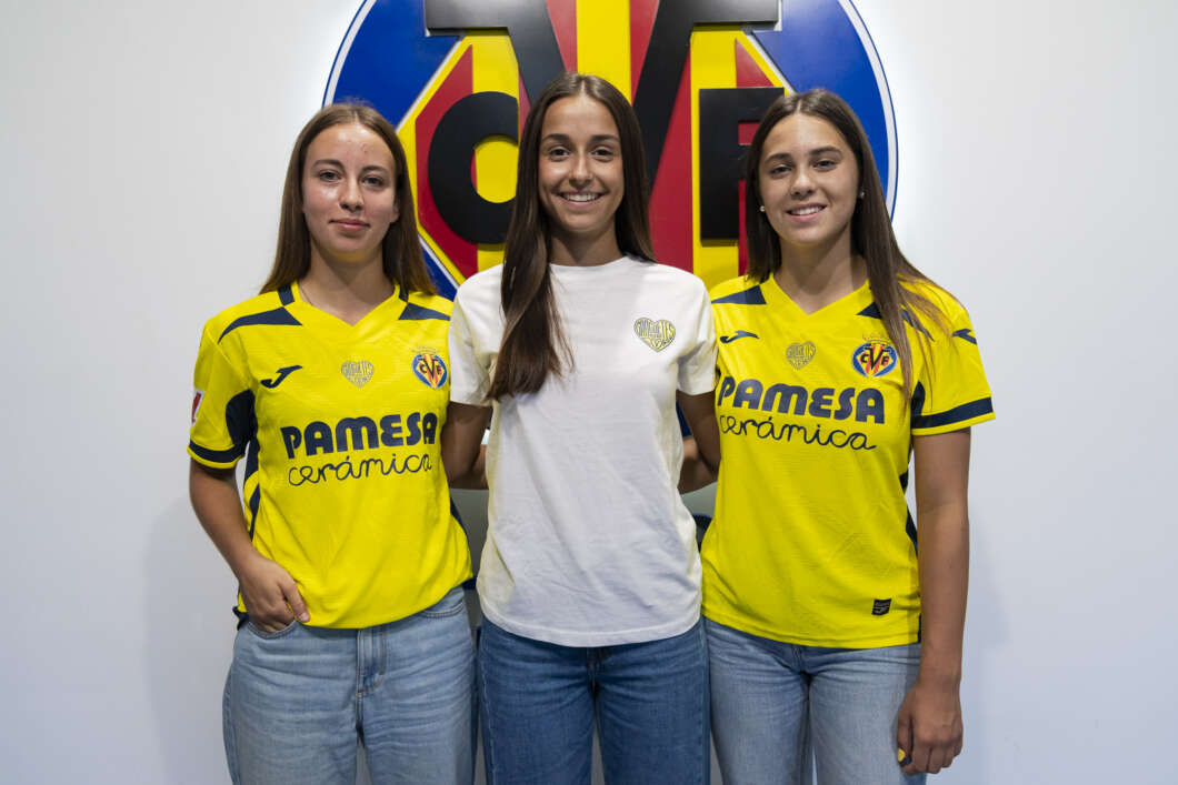 Three young women posing for a photo; two wear yellow Villarreal CF jerseys and one in a white T-shirt, with a large club emblem behind them.