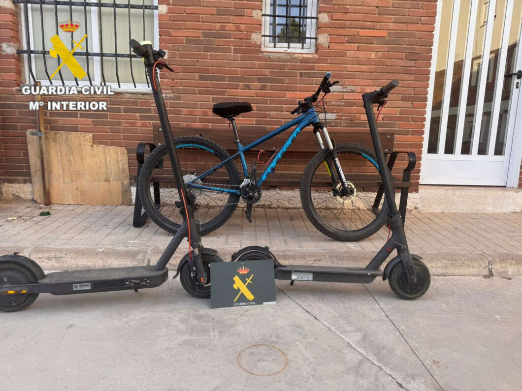 Two black electric scooters parked on a sidewalk with a blue bike mounted on a rack behind them, and a Guardia Civil emblem visible in the top-left corner of the image.
