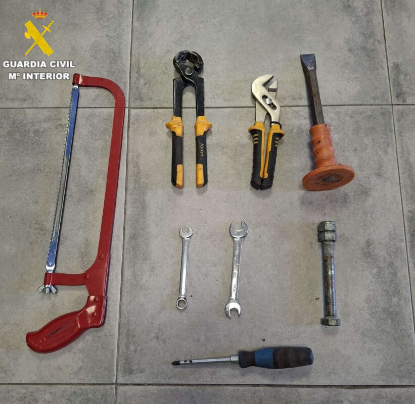 Tools laid out on a tiled floor: red hacksaw, yellow/black grips pliers, yellow-handled cutters, an orange-handled tool, two open-end wrenches, a large bolt, and a screwdriver, with a Guardia Civil logo in the corner.