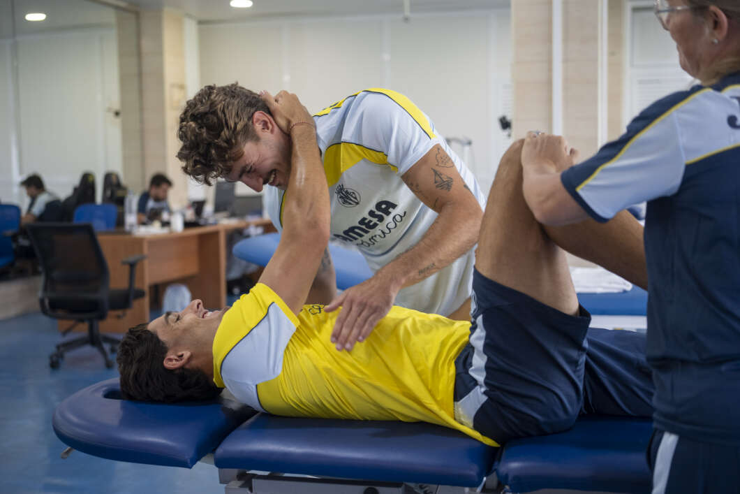 Athlete in a yellow shirt lying on a therapy table while a trainer assists by guiding his leg in a stretch in a rehab clinic.