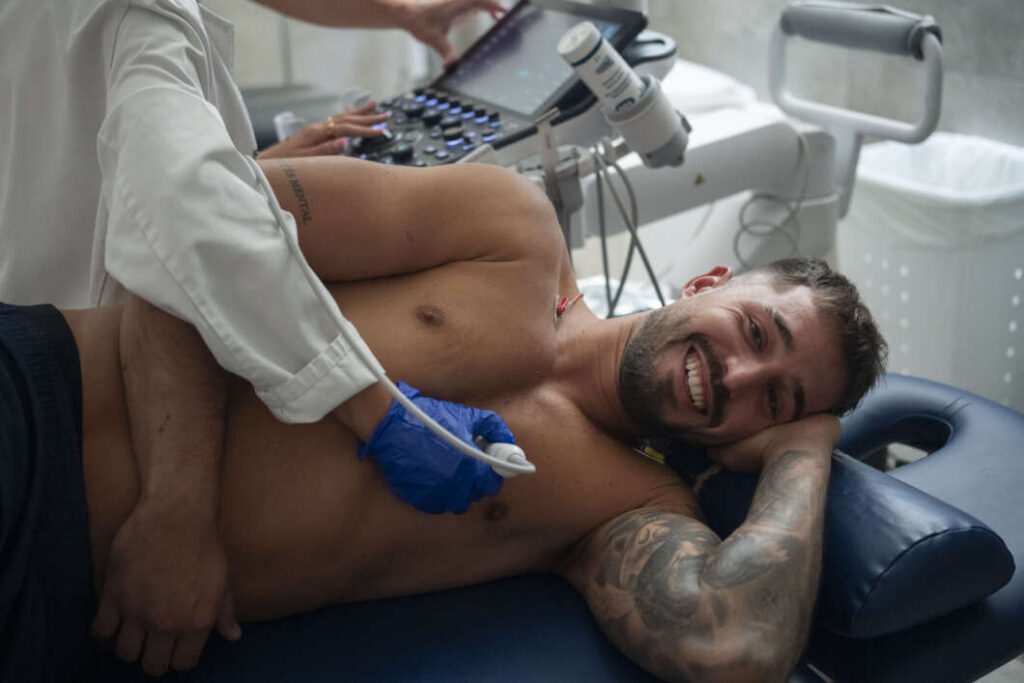 Smiling shirtless man lying on an examination table while a clinician uses an ultrasound probe on his chest on a medical device.