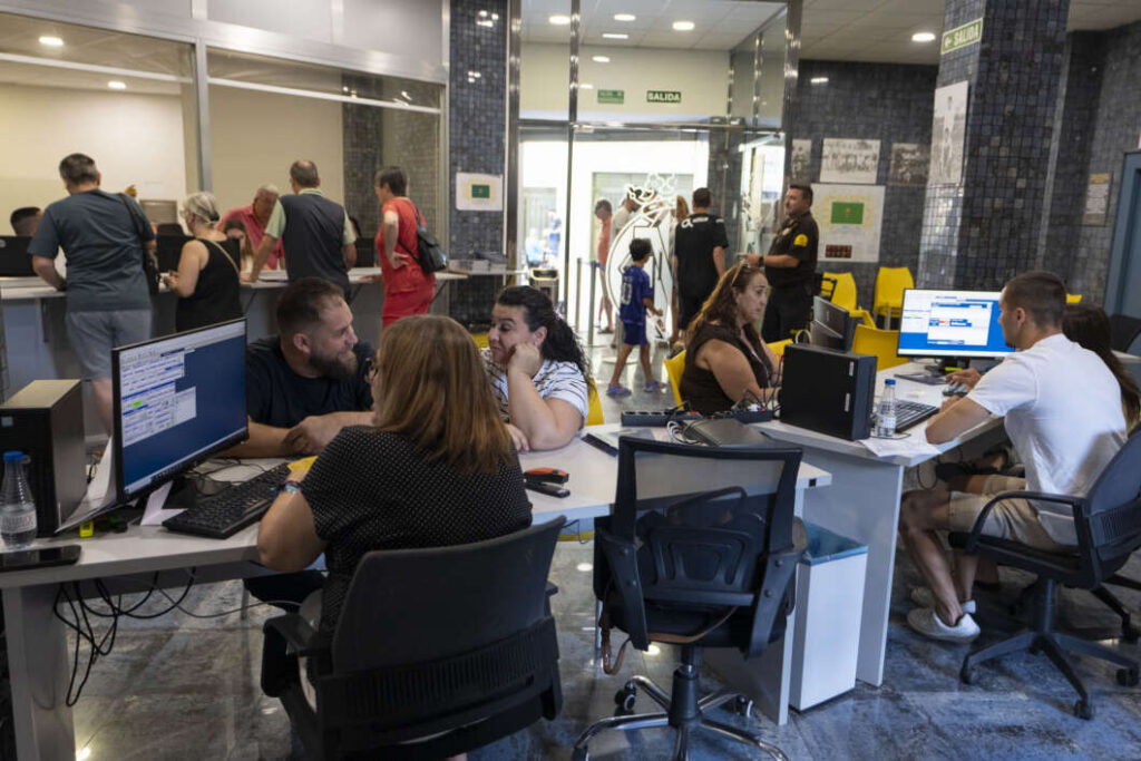 Busy reception area with staff at computer desks and customers at a counter; glass doors and an SALIDA sign in the background.