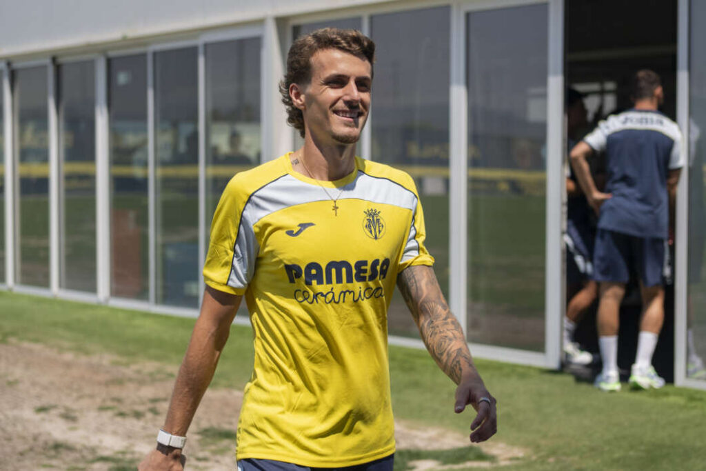 Smiling soccer player in a yellow training jersey walking outside a glass-fronted facility.