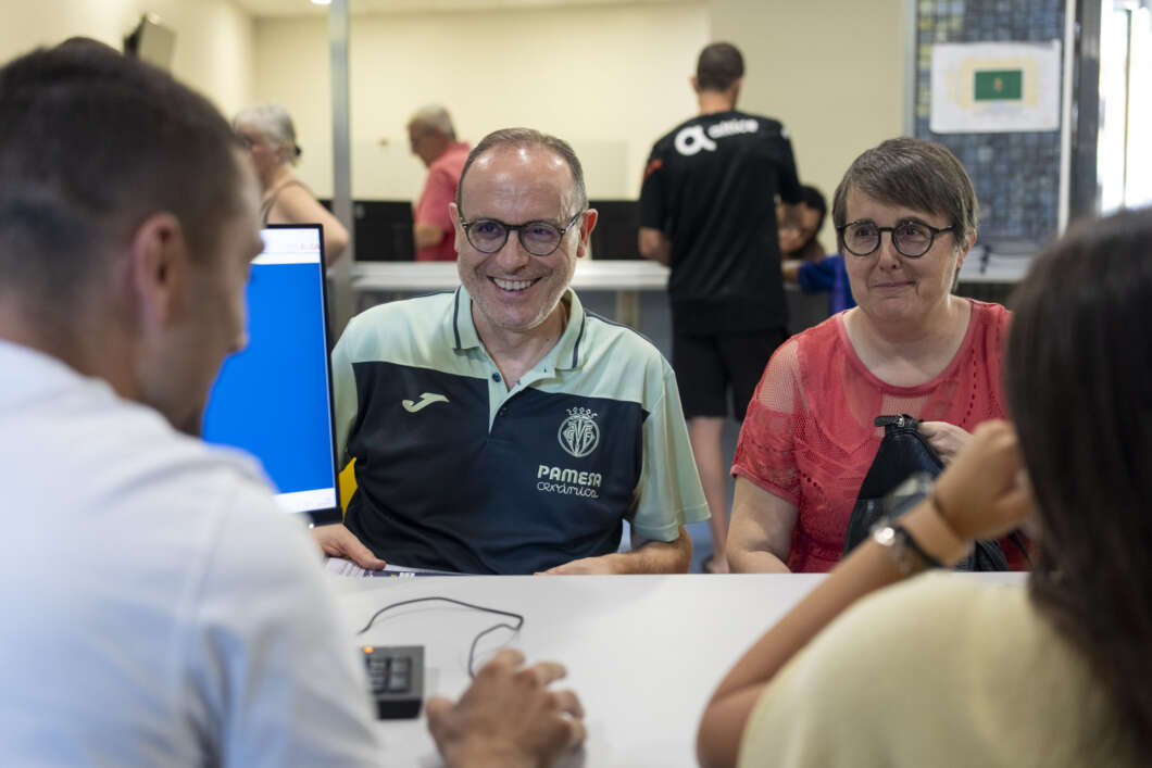 Smiling man in glasses wearing a polo with a crest sits at a service desk with a woman in a red top beside him, as a staff member assists them at a computer nearby.