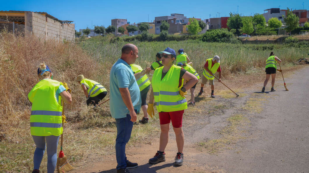 Group of volunteers in neon vests cleaning a roadside field with brooms and rakes.