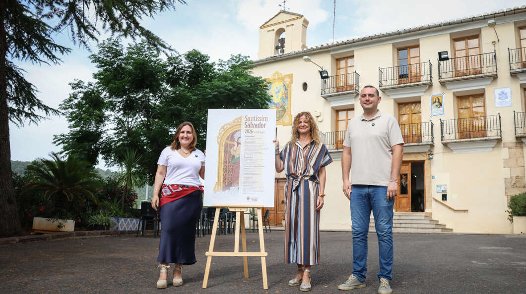 Three adults stand outdoors beside an easel with a tall poster, in front of a light-colored building with balconies and trees behind them.